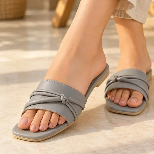 A person wearing light gray slide sandals with knotted front straps, shown from the calves down on a sunlit stone floor. The person is also wearing light beige linen pants.